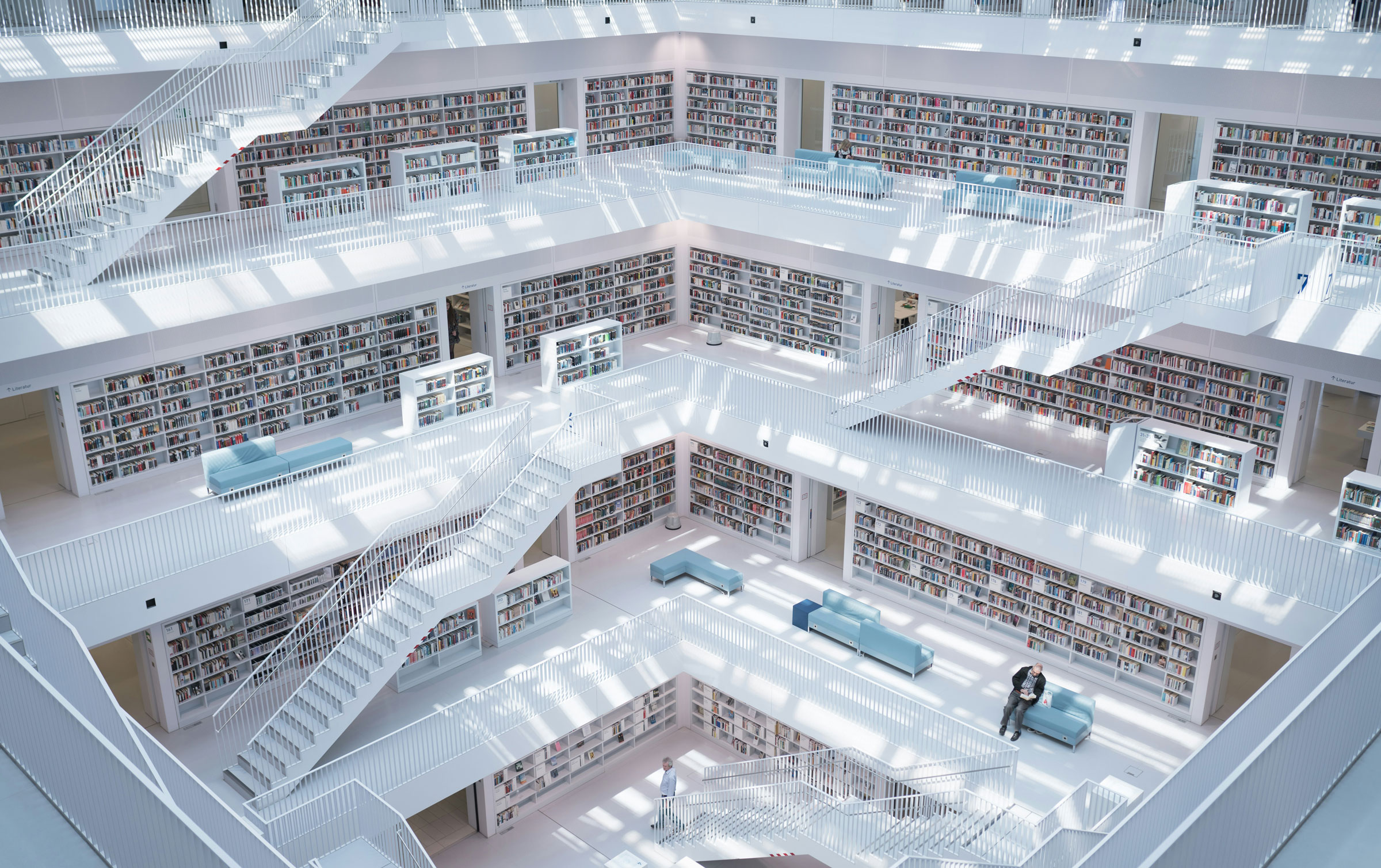 kdm-blog-header-1-0925 Innenansicht einer Bibliothek, die auf mehreren Ebenen mit weißen Treppen, mit Büchern gefüllten Regalen und Sitzgelegenheiten ausgestattet ist.